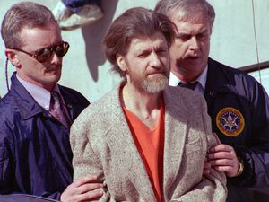(John Youngbear | The Associated Press) In this April 4, 1996 file photo Theodore John Kaczynski is flanked by federal agents as he is led to a car from the federal courthouse in Helena, Mont. A federal grand jury in San Francisco indicted Vladislav Victorvic Timoshchuk, on charges of mailing two envelopes containing ricin to Pelican Bay State Prison, officials announced on Friday Feb. 21, 2020. Last year the federal Bureau of Prisons intercepted a Christmas card from Timoshchuk, now living in Belarus, to Kaczynski, the convicted Unabomber, which authorizes say Timoshchuk discussed his plan to mail ricin to the United States. Kaczynski is serving a life sentence in a federal prison in Colorado for carrying out a series of mail bombings that killed three people.